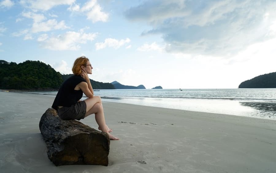 A woman sitting on driftwood, looking out at the sea